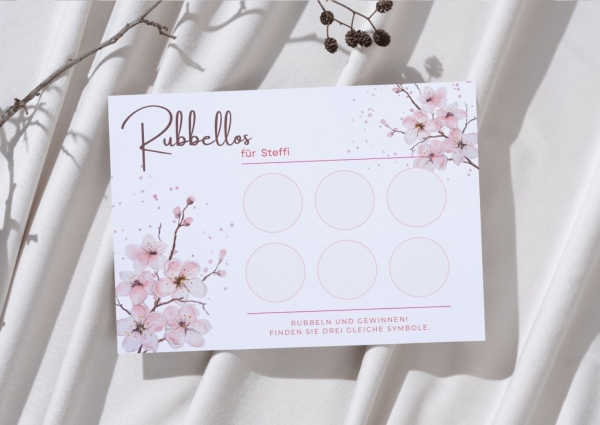 Personalisierte Rubbelkarte mit dem Namen „Steffi“ im zarten Blütendesign in Rosa. Die Karte liegt auf weichem, hellem Stoff mit Zweigen im Hintergrund. Sechs leere Rubbelfelder sind sichtbar.