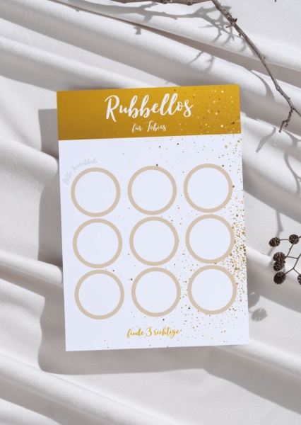 Rubbelkarte im gold-weißen Design mit dem Namen „Tobias“ und neun leeren Rubbelfeldern. Oben steht der Schriftzug „Rubbellos“, unten der Hinweis „Finde 3 richtige“. Die Karte liegt auf hellem Stoff mit dekorativen Zweigen.