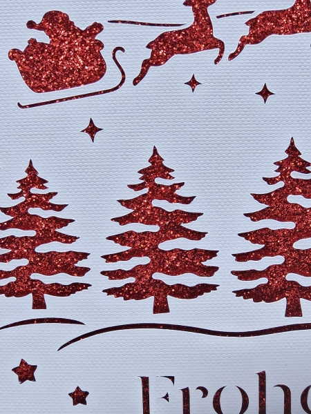 Detail des filigranen Designs einer handgefertigten Weihnachtskarte in Weiß mit rotem Glitzerhintergrund. Zeigt die glitzernden Weihnachtsbäume, Sterne und den Schlitten des Weihnachtsmanns, gezogen von Rentieren.