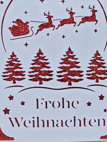 Detailansicht einer handgefertigten Weihnachtskarte in Weiß mit glitzerndem roten Hintergrund. Nahaufnahme des filigranen Designs mit Weihnachtsbäumen, Sternen, dem Schlitten des Weihnachtsmanns und dem ausgeschnittenen Schriftzug 'Frohe Weihnachten'.