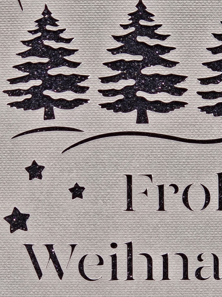 Detailansicht einer handgefertigten Weihnachtskarte in Grau mit schwarzem Glitzerhintergrund. Nahaufnahme zeigt glitzernde Weihnachtsbäume, filigran geschnittene Sterne und den Schriftzug 'Frohe Weihnachten'.