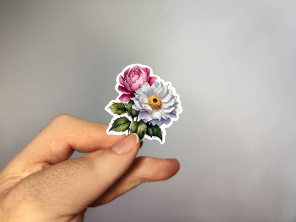 Hand, die einen Blumenaufkleber mit rosa und weißer Blüte sowie grünen Blättern hält, gestaltet von DreamArt VM.