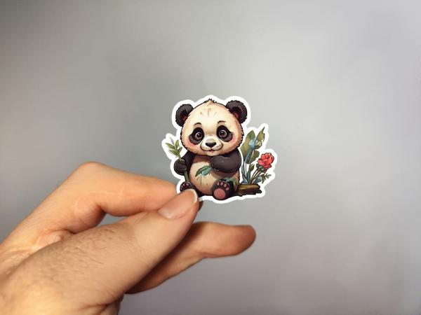 Hand, die einen Aufkleber in Form eines niedlichen Pandas mit Bambus und Blumen hält, gestaltet von DreamArt VM.