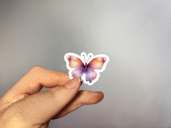 Hand, die einen Aufkleber in Form eines bunten Schmetterlings hält, gestaltet von DreamArt VM.
