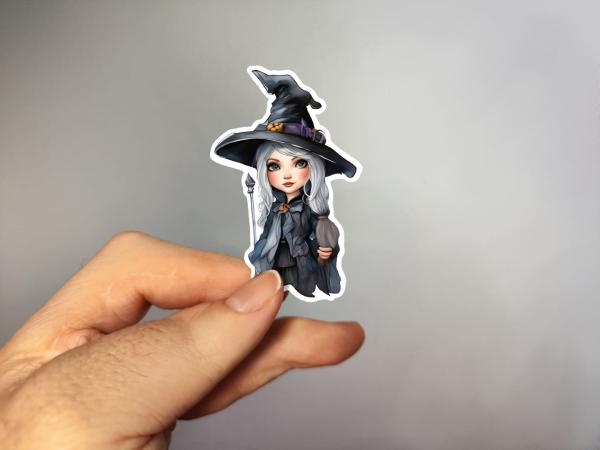 Hand, die einen Aufkleber in Form einer kleinen Hexe mit langen grauen Haaren, schwarzem Hut und schwarzem Kleid hält, gestaltet von DreamArt VM.