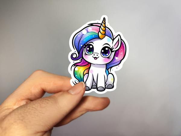 Hand, die einen Aufkleber in Form eines niedlichen Einhorns mit Regenbogenmähne und großen Augen hält, gestaltet von DreamArt VM