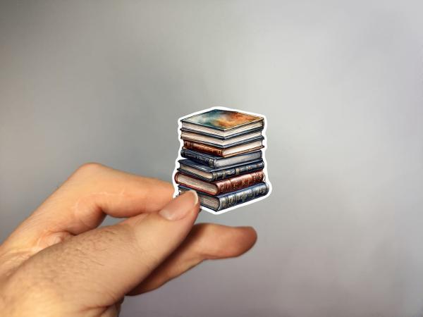 Hand hält einen Sticker mit einem Stapel alter Bücher in verschiedenen Farben, die sorgfältig übereinander gestapelt sind.