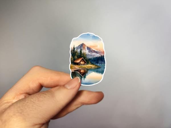 Ein Aufkleber mit einem malerischen Bergsee bei Sonnenuntergang, umgeben von hohen Tannen. Im Vordergrund steht eine gemütliche Hütte am Seeufer, die sich im klaren Wasser spiegelt. Im Hintergrund erhebt sich ein majestätischer Berg, der in sanftes, warme