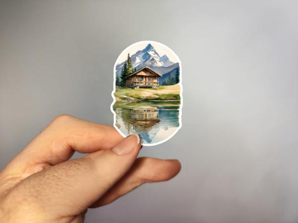 Handgefertigter Aquarell-Aufkleber mit Berghütte und Seespiegelung – in der Hand gehalten
