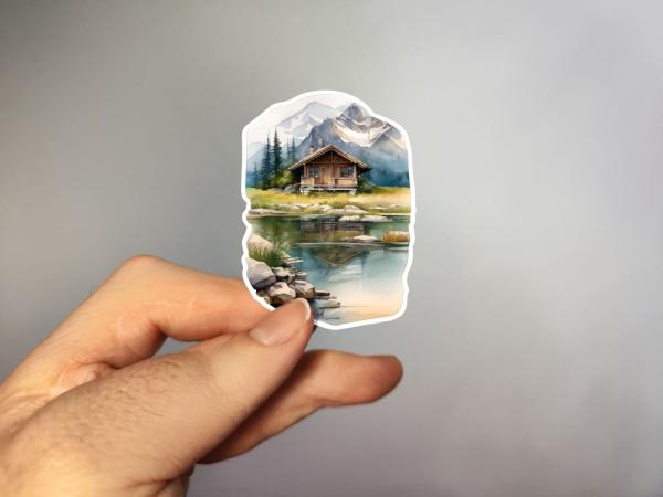 Handgefertigter Aquarell-Aufkleber mit Berghütte und Seespiegelung – in der Hand gezeigt