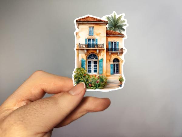 Ein handgehaltener Sticker, der eine mediterrane Villa mit hellgelber Fassade, blauen Fensterläden, einem Balkon und einem großen Bogenfenster zeigt. Die Villa ist umgeben von grünen Pflanzen, die dem Gebäude ein lebendiges und exotisches Flair verleihen.