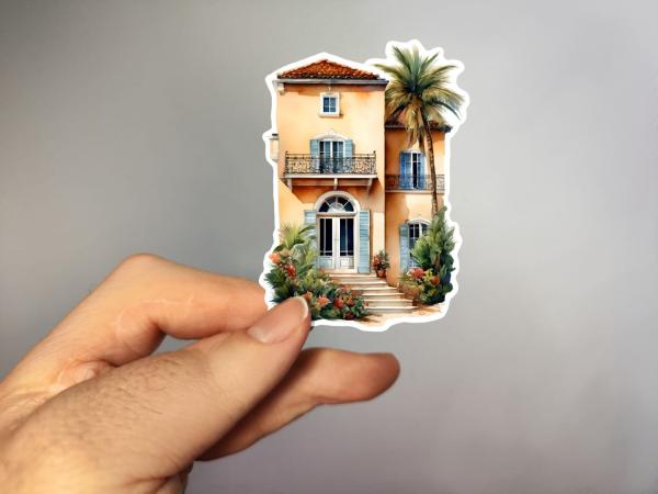 Ein handgehaltener Sticker, der eine elegante mediterrane Villa mit hellgelber Fassade, blauen Fensterläden und einem Balkon zeigt. Vor der Villa befindet sich eine Treppe, die von tropischen Pflanzen und Palmen umgeben ist, was dem Gebäude einen exotisch