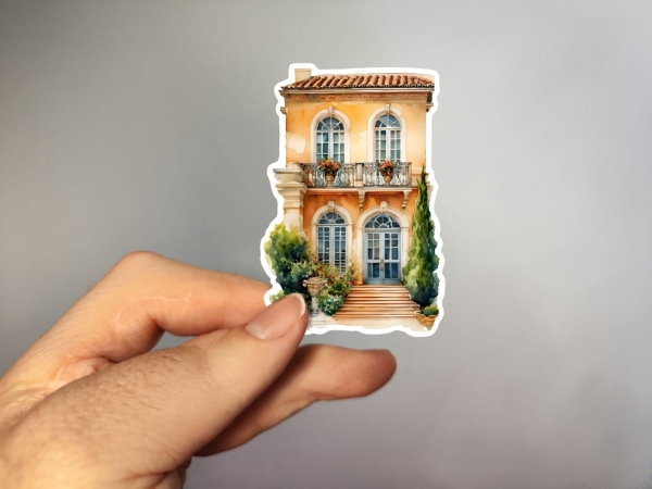 Aufkleber einer mediterranen Villa mit zwei Etagen, geschwungenen Balkontüren und dekorativem Balkon, flankiert von Zypressen und grünen Büschen. Bild D.