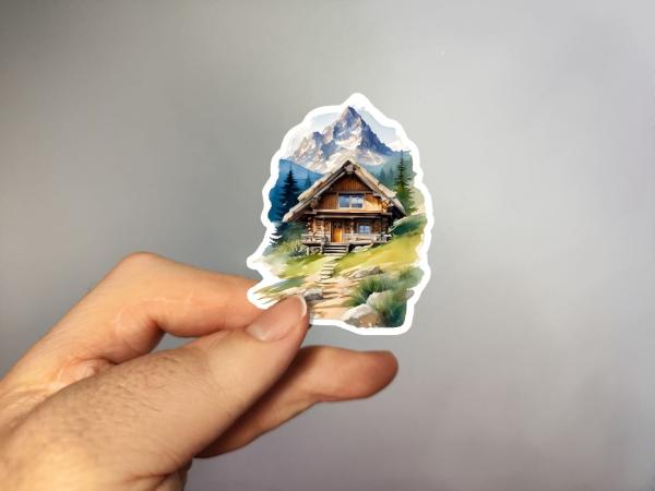 Sticker einer rustikalen Berghütte inmitten einer malerischen Alpenlandschaft mit einem schneebedeckten Berg im Hintergrund, umgeben von grünen Wiesen und Nadelbäumen.