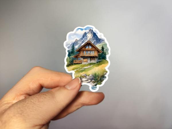 Sticker einer gemütlichen Berghütte auf einer grünen Wiese, umgeben von Bergen mit schneebedeckten Gipfeln im Hintergrund.