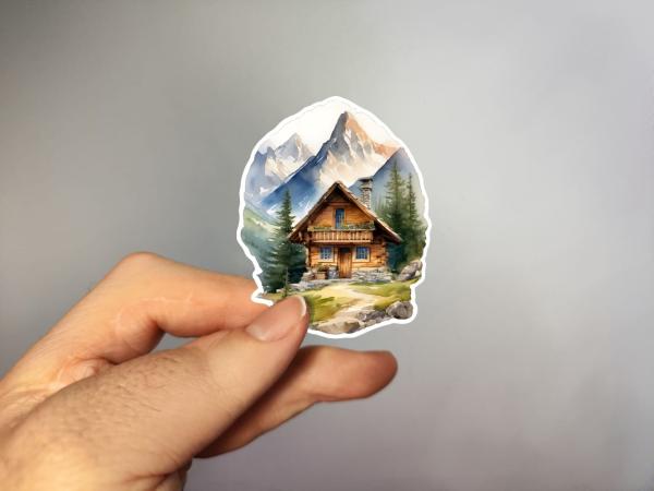 Sticker einer rustikalen Berghütte, eingebettet in eine Berglandschaft mit schneebedeckten Gipfeln und grünen Tannen im Hintergrund.