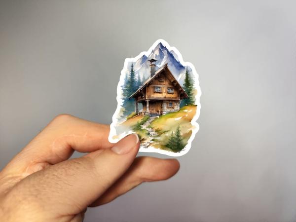 Sticker einer traditionellen Holzhütte in den Bergen, umgeben von grünen Bäumen und einem klaren Pfad, der zur Hütte führt, mit schneebedeckten Berggipfeln im Hintergrund.