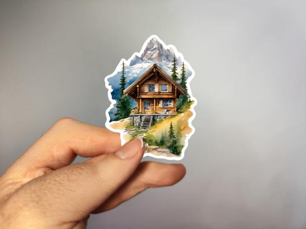 Sticker einer rustikalen Holzhütte in den Alpen, umgeben von grünen Bäumen, mit einem steinigen Pfad und schneebedeckten Berggipfeln im Hintergrund.
