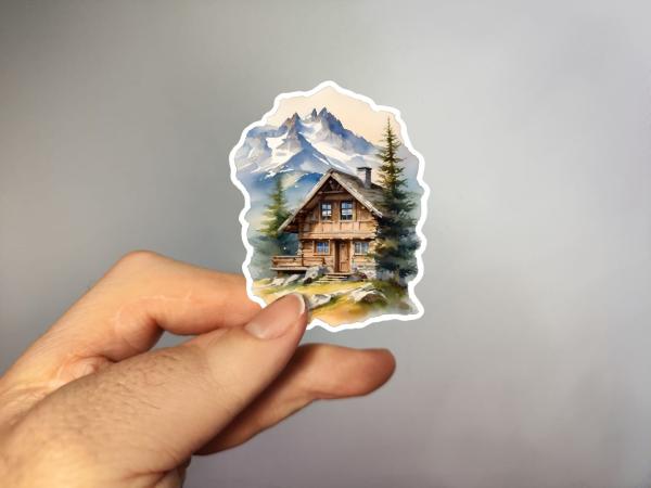 Sticker einer traditionellen Berghütte aus Holz in den Alpen, umgeben von Tannenbäumen und majestätischen Berggipfeln im Hintergrund.