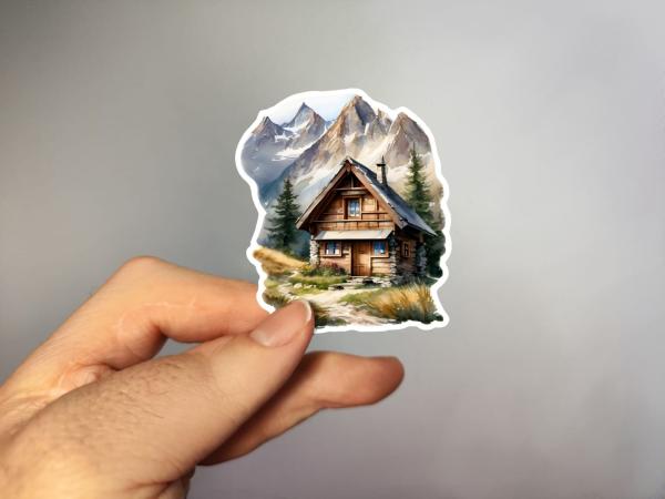 Sticker einer rustikalen Berghütte mit Steinfundament, umgeben von alpinen Wiesen und hohen Berggipfeln im Hintergrund.