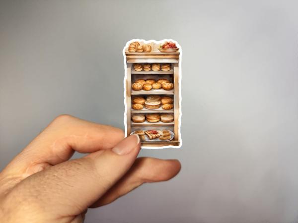 Hand hält einen Aufkleber, der eine Vitrine mit verschiedenen Backwaren zeigt, Bild D. Die Illustration zeigt eine reichhaltige Auswahl an Brötchen, Broten und Gebäck, die auf mehreren Regalen gestapelt sind, mit einer Obstschale auf dem obersten Regal.
