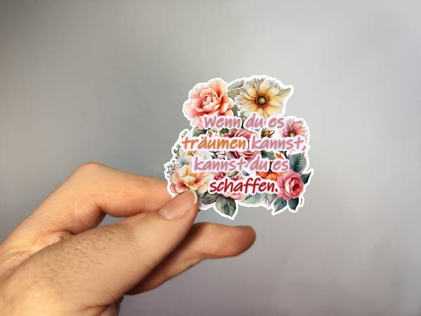 Hand, die einen Blumenaufkleber mit dem Text 'Wenn du es träumen kannst, kannst du es schaffen.' in Rosa und Orange hält, gestaltet von DreamArt VM.