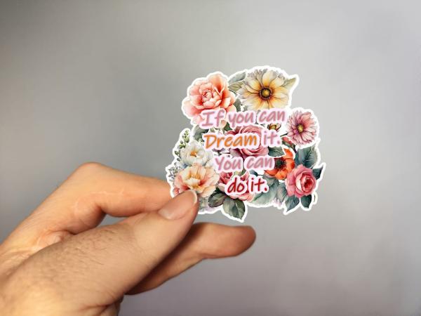 Hand holding a floral sticker with the motivational text 'If you can dream it, you can do it.' in pink and orange, designed by DreamArt VM.