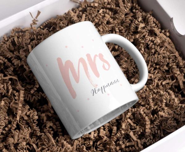 Weißer Keramikbecher in einer Geschenkbox mit braunem Papierfüllmaterial, bedruckt mit dem Wort 'Mrs' in einer großen, rosa Schrift und dem Namen 'Hoffmann' darunter in einer eleganten schwarzen Schrift. Der Becher ist schräg platziert, sodass der Druck g