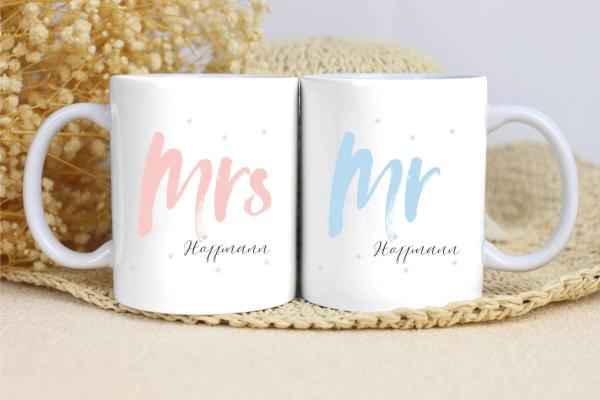 Zwei weiße Keramikbecher nebeneinander auf einer beigen Unterlage, umgeben von Trockenblumen im Hintergrund. Der linke Becher trägt die Aufschrift 'Mrs' in einer großen, rosa Schrift, und darunter den Namen 'Hoffmann' in einer eleganten schwarzen Schrift.