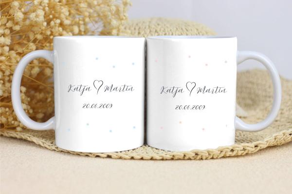 Zwei weiße Keramikbecher nebeneinander auf einer beigen Unterlage, umgeben von Trockenblumen im Hintergrund. Beide Becher zeigen den Text 'Katja  Martin' in eleganter Schrift, darunter das Datum '20.06.2009'. Die linke Tasse ist mit blauen Punkten dekorie