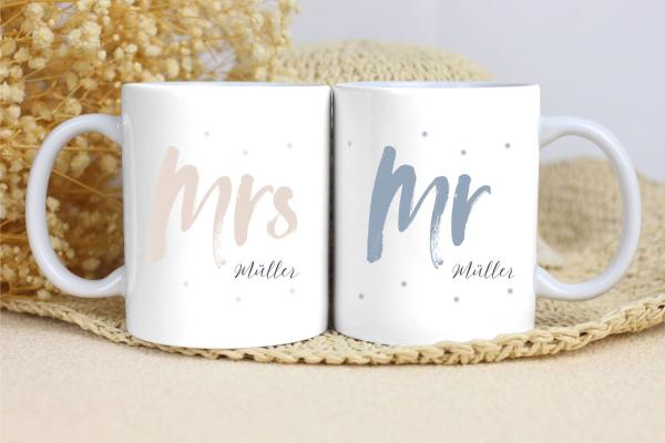 Zwei weiße Keramikbecher nebeneinander auf einer beigen Unterlage mit Trockenblumen im Hintergrund. Der linke Becher zeigt den Text 'Mrs Müller' in einem sanften Beigeton mit kleinen beigefarbenen Punkten als Verzierung. Der rechte Becher zeigt den Text '