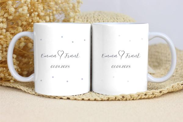 Zwei weiße Keramikbecher nebeneinander auf einer beigen Unterlage mit Trockenblumen im Hintergrund. Beide Becher zeigen den Text 'Emma  Frank' mit dem Datum '08.04.2023' darunter. Der linke Becher hat kleine blaue Punkte als Verzierung, während der rechte