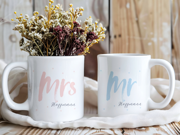 Zwei personalisierte Tassen mit den Aufschriften „Mrs Hoffmann“ in Rosa und „Mr Hoffmann“ in Hellblau, verziert mit feinen Punkten. Romantisches Geschenk für Paare, zur Hochzeit oder zum Valentinstag.
