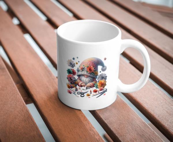 Weißer Kaffeebecher mit niedlichem Gnom-Design, umgeben von bunten Blumen und Bastelutensilien, auf einer Holzoberfläche. Perfektes Geschenk für kreative Seelen und Gartenliebhaber.