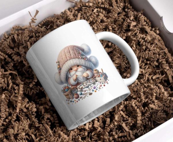 Weiße Tasse mit einem niedlichen Weihnachtszwerg-Motiv, der in einer gemütlichen Wintermütze und Schal gekleidet ist und von verschiedenen Bastelmaterialien umgeben ist, wie Perlen, Garn und Pinsel. Die Tasse liegt in einer Geschenkbox, die mit braunem Pa
