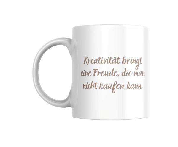 Ein weißer Keramikbecher mit dem Zitat 'Kreativität bringt eine Freude, die man nicht kaufen kann' in eleganter Schriftart. Der Becher steht auf einem neutralen Hintergrund.