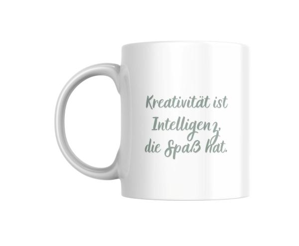 Ein weißer Keramikbecher steht auf einem einfarbigen Hintergrund. Der Becher ist mit einer inspirierenden Aufschrift verziert, die lautet: 'Kreativität ist Intelligenz, die Spaß hat.' Die Schrift ist in einem sanften Grünton gehalten und verleiht dem Bech