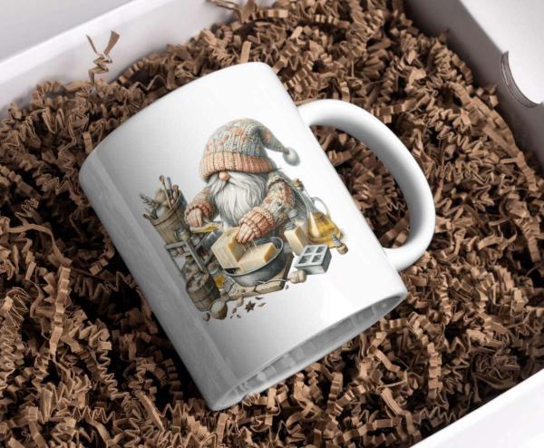 Weißer Keramikbecher mit einem Motiv eines kleinen Gnoms in Strickmütze und Pullover, der in einer Werkstatt an der Herstellung von handgemachter Seife arbeitet. Der Becher liegt in einer Geschenkbox auf braunem Füllmaterial und ist ideal für Liebhaber vo