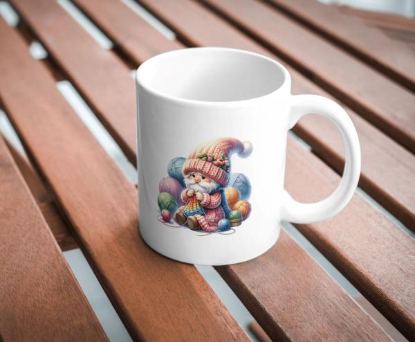 Weißer Keramikbecher mit einem Motiv eines niedlichen Gnoms in einer bunten Strickmütze, der mit farbenfrohen Garnknäueln strickt. Der Becher steht auf einem Holztisch und ist perfekt für Handarbeitsliebhaber und kreative Menschen.