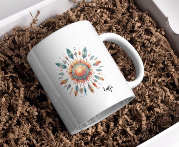Weißer Keramikbecher mit personalisiertem Namen 'Katja', verziert mit einem Boho-Sonnenmotiv in warmen Erdtönen und türkisfarbenen Akzenten, umgeben von Federn und spirituellen Symbolen. Der Becher ist in einer Geschenkbox mit dekorativem Papier verpackt,