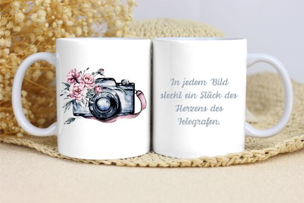 Weißer Keramikbecher im Set mit zwei Seiten: Die linke Seite zeigt eine nostalgische Kamera im Aquarellstil mit rosa Blumen, die die Linse umrahmen. Auf der rechten Seite ist der Schriftzug 'In jedem Bild steckt ein Stück des Herzens des Fotografen' in el