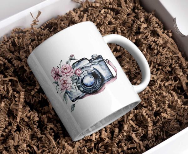 Weißer Keramikbecher in Geschenkverpackung mit einer Aquarellillustration einer Vintage-Kamera mit rosa Blüten, die sich um das Objektiv ranken. Der Becher liegt in einer naturfarbenen Papierpolsterung und verleiht dem Produkt eine rustikale und elegante