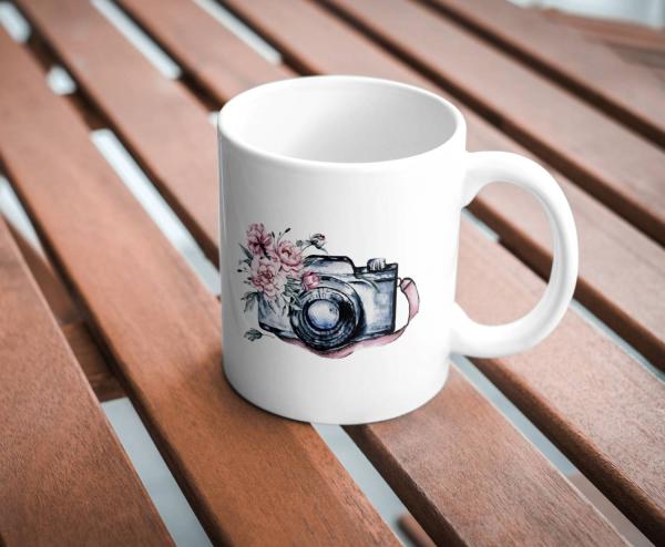 Weißer Keramikbecher mit einer Aquarellillustration einer Vintage-Kamera, die mit rosa Blumen verziert ist. Der Becher steht auf einem Holztisch im Freien und vermittelt eine rustikale, natürliche Atmosphäre.