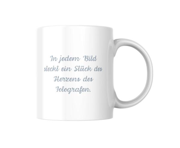 Weißer Keramikbecher mit einem Zitat in hellblauer, handgeschriebener Schrift: ‚In jedem Bild steckt ein Stück des Herzens des Fotografen.‘ Der Becher hat ein minimalistisches Design, das die Bedeutung des Zitats hervorhebt.