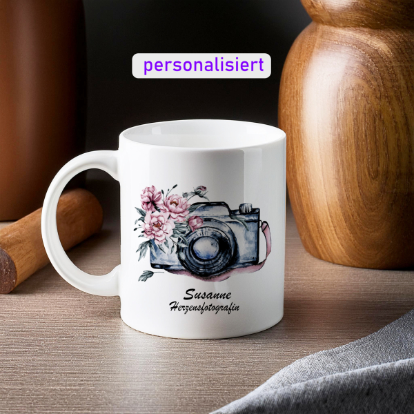 Weißer Keramikbecher mit einer Aquarellillustration einer Vintage-Kamera, geschmückt mit rosa Blüten, und dem Schriftzug 'Veronika Joy Fine Art Photography & Film'. Der Becher ist festlich arrangiert mit einem Zweig Tannenzweigen und goldenen Weihnachtsku