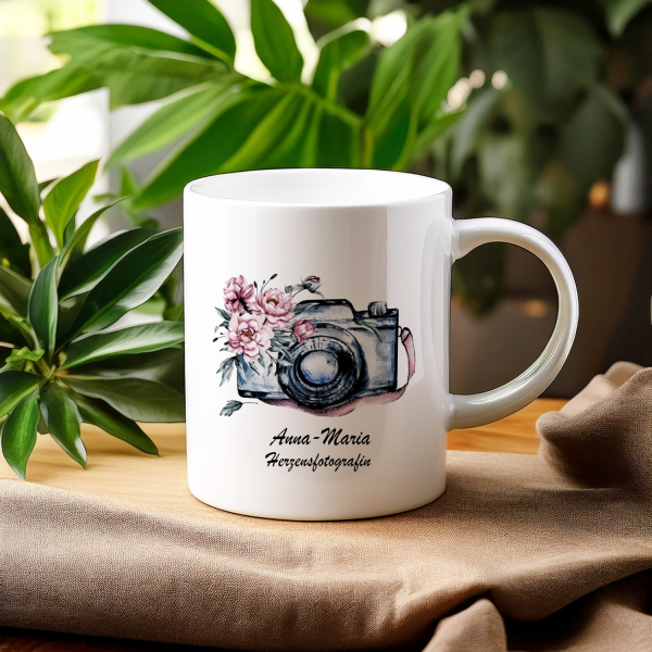 Weißer Keramikbecher mit einer Aquarellillustration einer Vintage-Kamera, umgeben von rosa Blüten, und dem Namen 'Susanne' darunter. Der Becher ist festlich arrangiert mit einem Zweig Tannenzweigen und goldenen Weihnachtskugeln im Hintergrund, was eine el