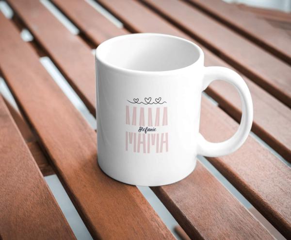 Weiße Tasse mit dem Schriftzug „MAMA“ in zartem Rosaton und dem Namen „Stefanie“ darunter in dunkler Schrift, verziert mit drei kleinen Herzen darüber. Die Tasse steht auf einem hölzernen Tisch und das Design ist schlicht und elegant, ideal als persönlich