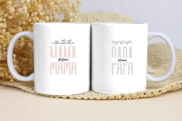 Zwei weiße Tassen mit schlichtem Design. Die linke Tasse trägt den Text „MAMA“ in einem zarten Rosaton mit dem Namen „Stefanie“ darunter und drei kleinen Herzen darüber. Die rechte Tasse zeigt den Text „PAPA“ in Grau mit dem Namen „Johann“ darunter und kl
