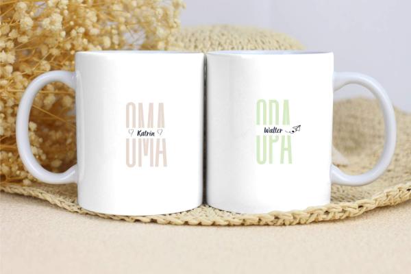 Zwei weiße Tassen mit schlichten Designs. Die linke Tasse zeigt den Text „OMA“ in einem weichen Beige, mit dem Namen „Katrin“ darunter und zwei kleinen Herzen auf beiden Seiten. Die rechte Tasse trägt den Text „OPA“ in einem zarten Grün, mit dem Namen „Wa