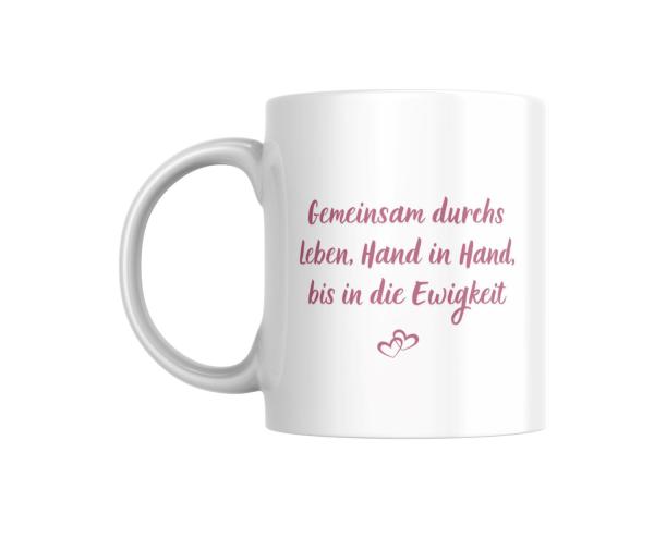 Weiße Tasse mit romantischem Spruch in rosaner Schrift: ‚Gemeinsam durchs Leben, Hand in Hand, bis in die Ewigkeit‘ und zwei ineinandergreifenden Herzen darunter. Ein liebevolles Design, ideal als Geschenk für Paare.